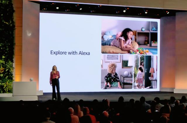 Photo from an Amazon launch event. A presenter stands on stage in front of a huge screen. A slide reads "Explore with Alexa" with a collage of images. Packed audience.