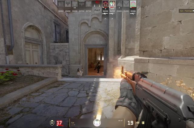 Gameplay still from Counter-Strike 2. The player (first-person view) holds an assault rifle as they head down an outdoor corridor of historical buildings. An enemy is being shot in the player's reticle.
