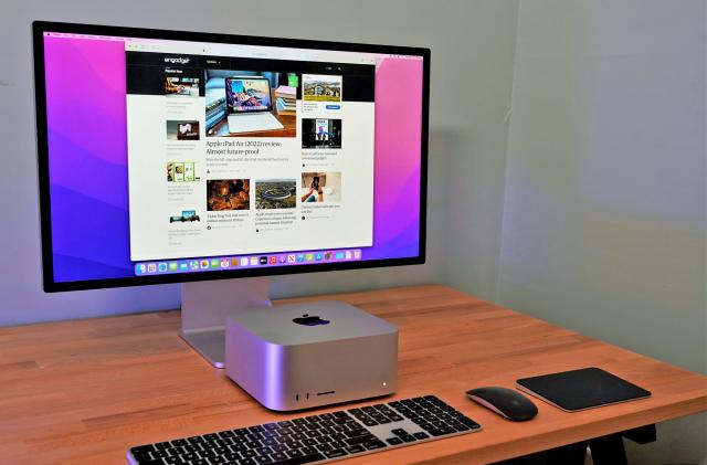 A sparse desktop setup is shown with a Apple Mac Studio and Studio Display sitting on a wooden desktop alongside a keyboard, mouse and touchpage.