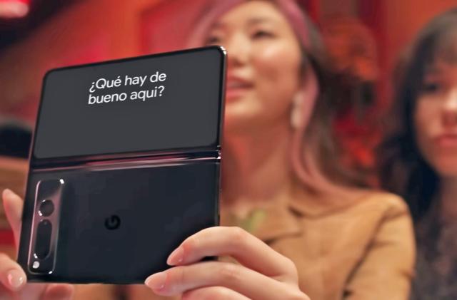 Still from a Google promotional video, featuring two women in a restaurant booth holding up a Pixel Fold phone (the phone is in focus, the women are blurred). The phone's outer screen shows the translation of her spoken words: "¿Qué hay de bueno aqui?"