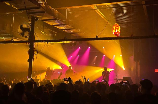 A band called Thrice is seen playing on stage from the perspective of an audience member. People are holding up phones to film them and the band is lit by pink and yellow spotlights.