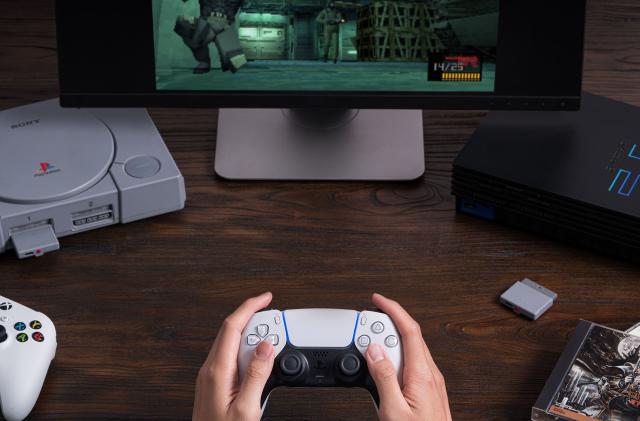 A pair of hands holds a PlayStation 5 DualSense controller. A PlayStation 1 and PlayStation 2 are in the background and a dongle sits on the table. A game is shown on a monitor.