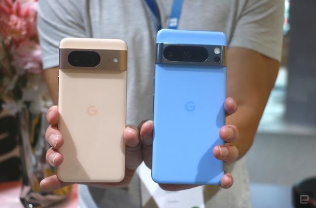 A person holding up the peach Pixel 8 and the blue Pixel 8 Pro with their rear cameras facing out.