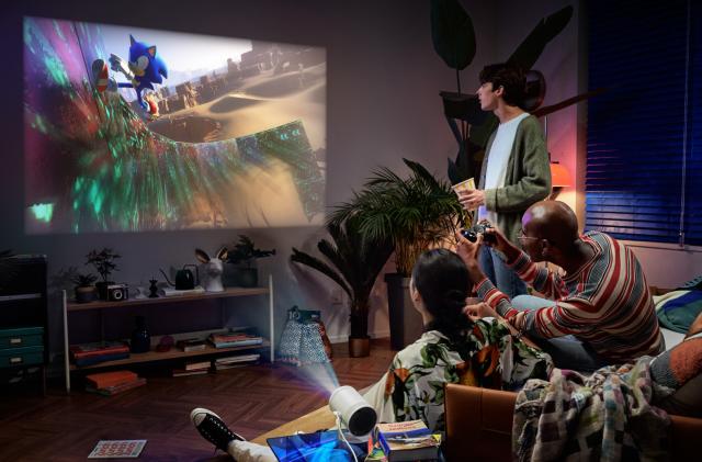 Three people in the living room, one holding a game controller, with the Samsung projector showing an image of Sonic the Hedgehog on the wall. 